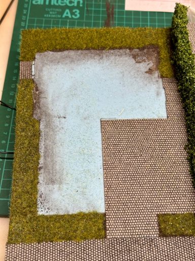 Modellbau-Landschaft mit Wasserfläche, Gras und gemischtem Untergrund.